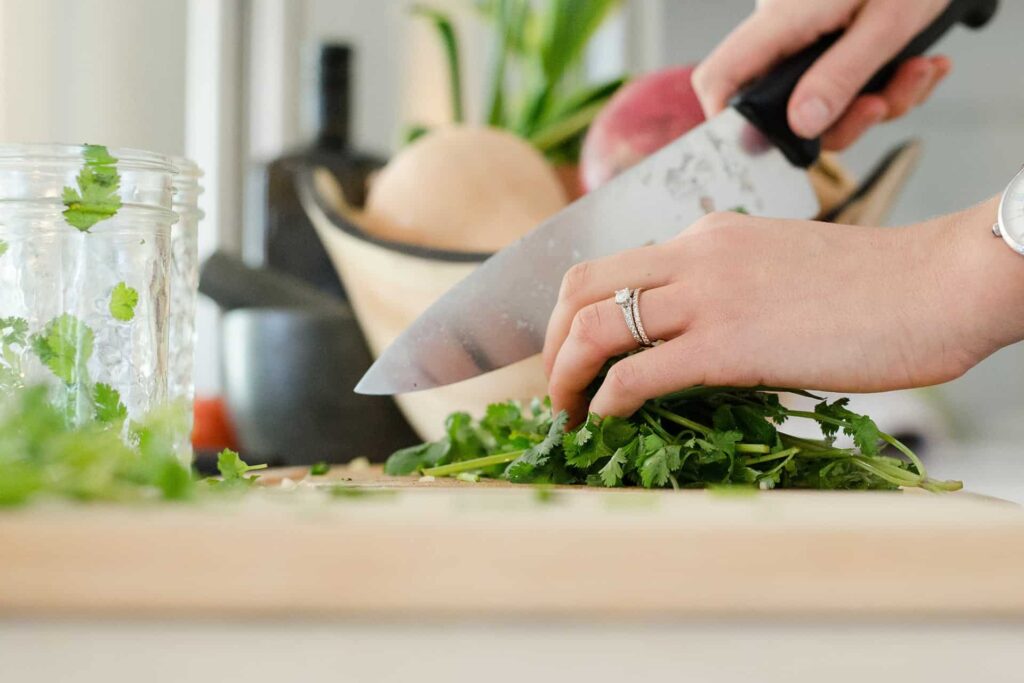 alyson-mcphee-yWG-ndhxvqY-unsplash - RIM Digital Close-up of hands chopping fresh cilantro with a large knife on a wooden cutting board, with a blender jar in the background.