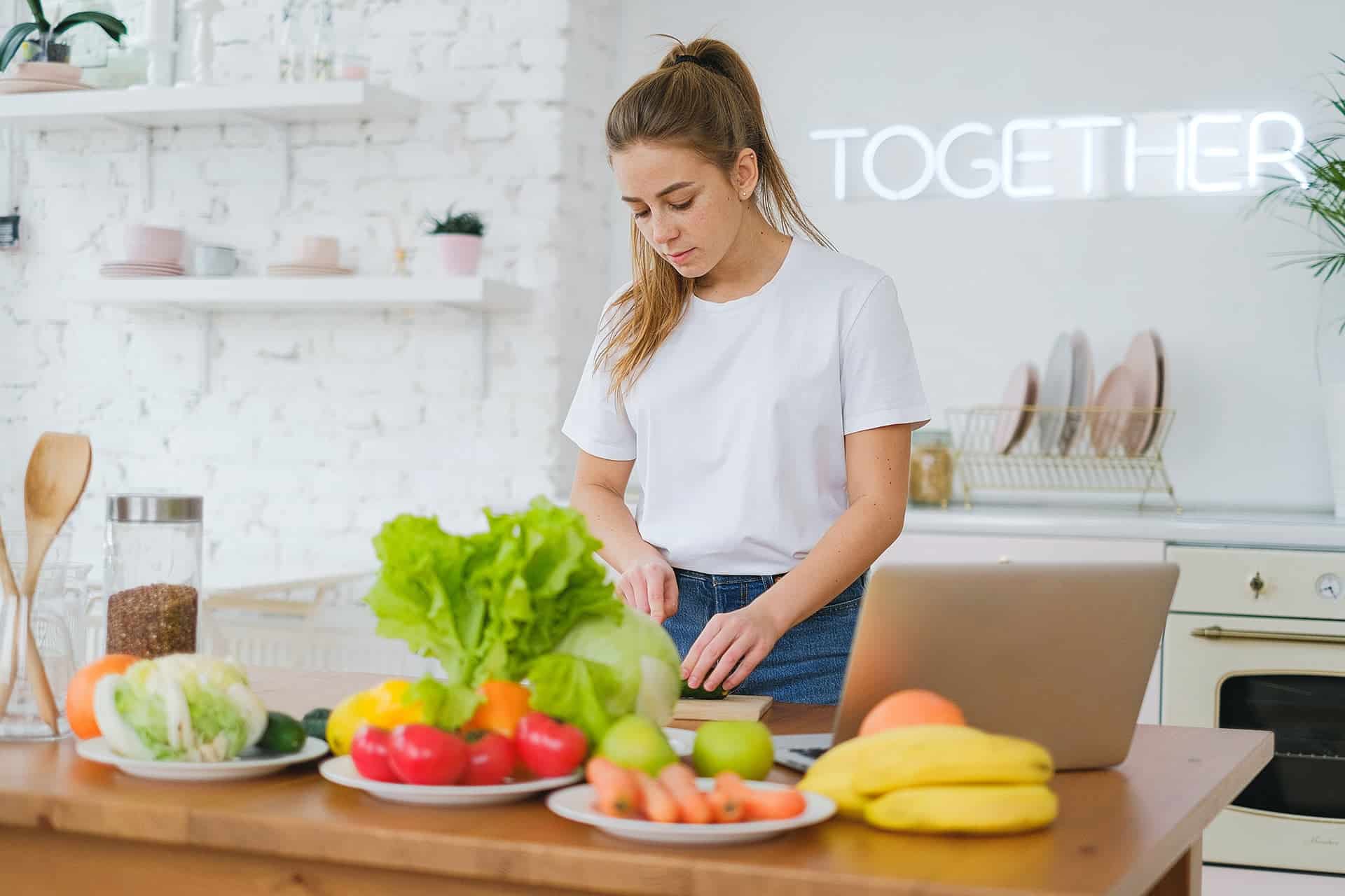 pexels-anna-shvets-6593581 - RIM Digital Woman in a white shirt chopping vegetables at a bright kitchen island; fresh produce and a laptop nearby, with a neon 'TOGETHER' sign in the background.