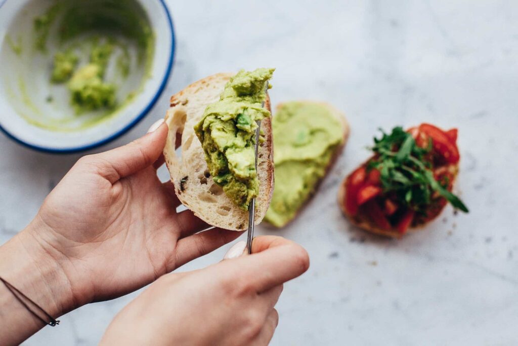 pexels-daria-shevtsova-1580467 - RIM Digital Close-up of a hand holding a bread slice with avocado spread, a fork lifting a chunk of avocado.