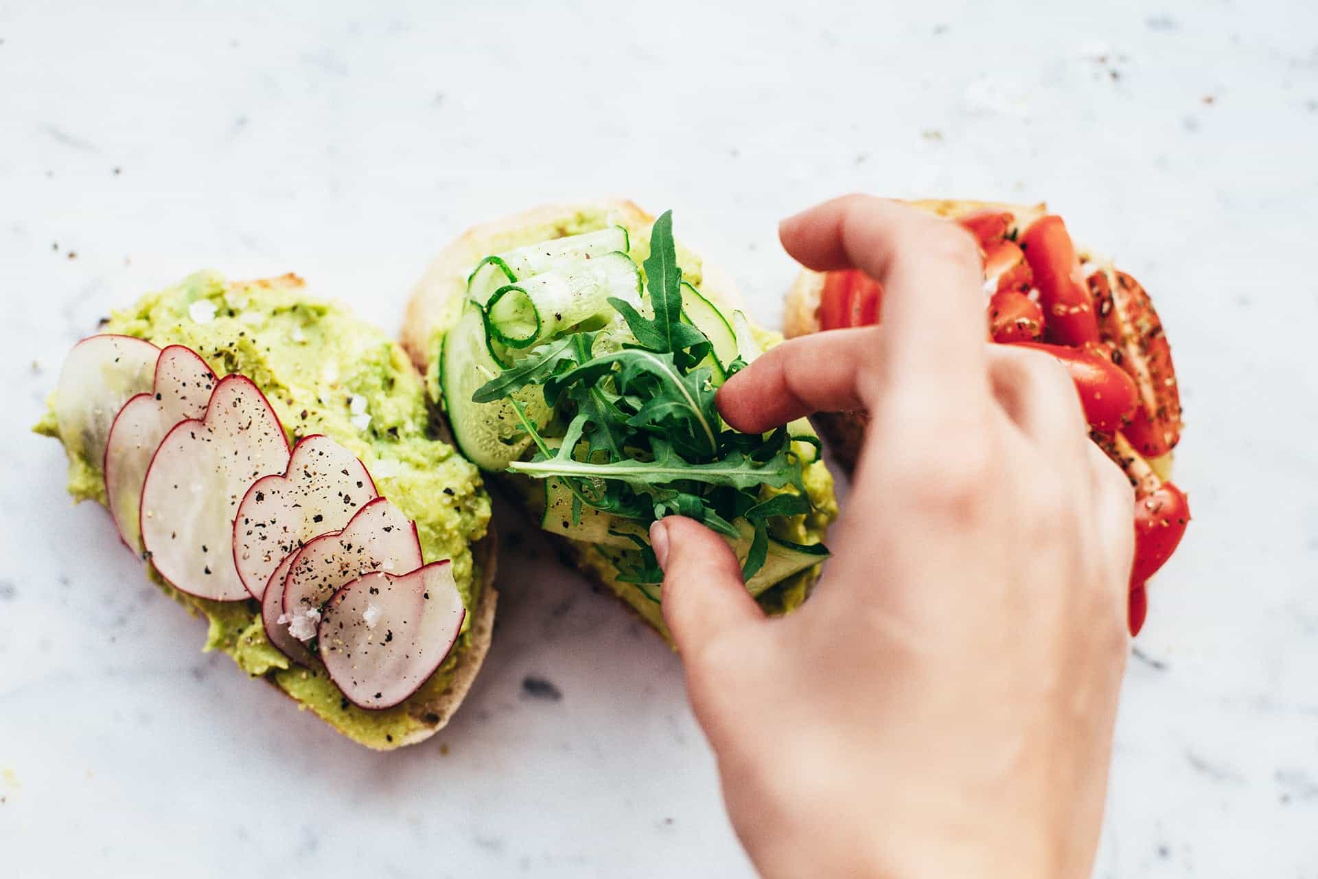 Hands assembling a colorful open-faced veggie sandwich with avocado spread, radish slices, cucumber ribbons, greens, and tomato on a marble surface.