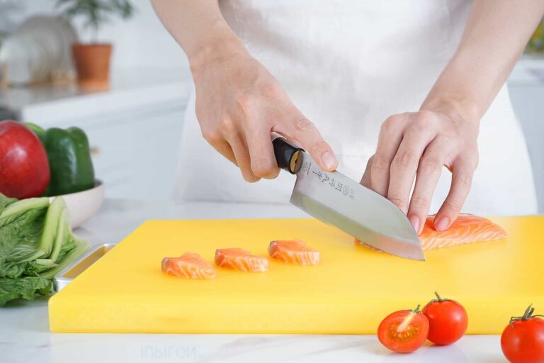 pexels-karolina-grabowska-666020312 - RIM Digital Person chopping salmon fillet into small pieces on a bright yellow cutting board with fresh vegetables nearby (greens and tomatoes) in a kitchen setting