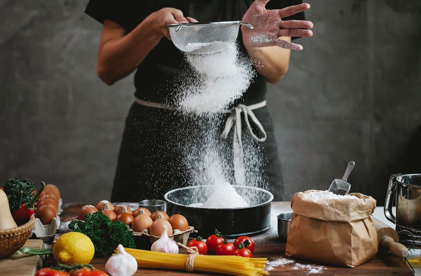 pexels-klaus-nielsen-6287225-2 - RIM Digital Person in a black shirt and apron sifts flour into a mixing bowl as flour falls midair in a kitchen scene.