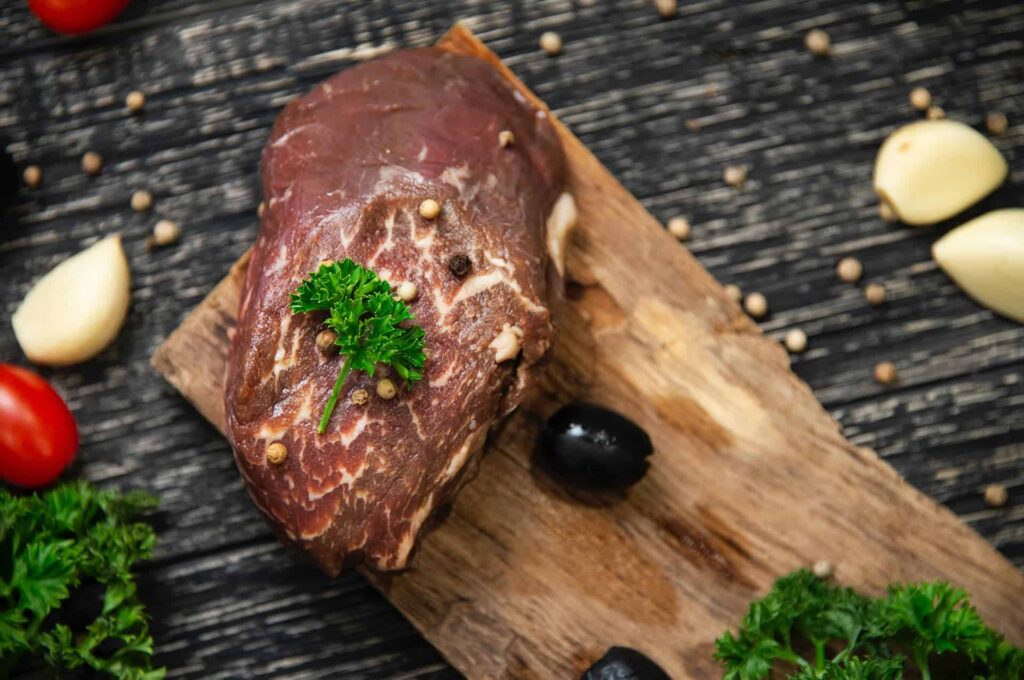 pexels-kristya-nugraha-5045088 - RIM Digital Raw beef steak resting on a wooden cutting board with parsley, garlic, and scattered peppercorns as seasoning.