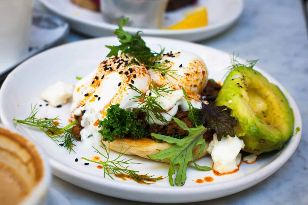 Toast topped with poached eggs, dill and herbs, sesame seeds, and chili oil, with avocado and greens on a white plate.