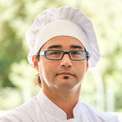 pexels-miquel-ferran-gomez-figueroa-3814446 - RIM Digital Close-up of a male chef wearing a white chef's hat and coat, with blue glasses, looking at the camera.