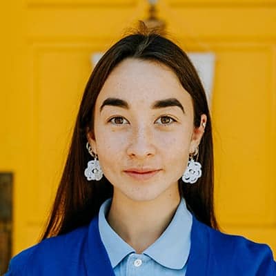 Portrait of a young woman with long dark hair wearing a blue blouse and large white floral earrings, in front of a bright yellow background.