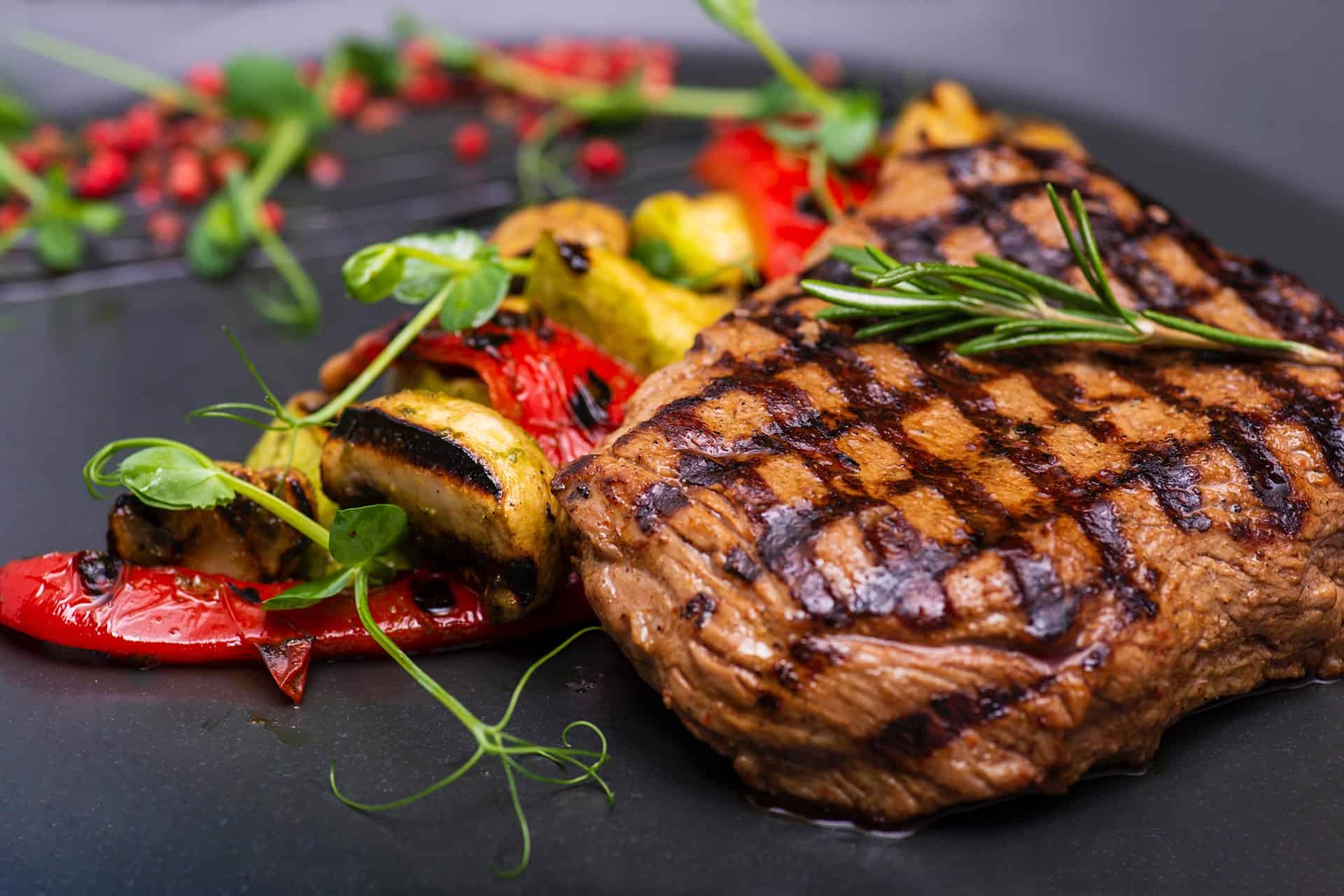 Juicy grilled steak topped with a rosemary sprig, served with roasted peppers and vegetables on a dark plate.