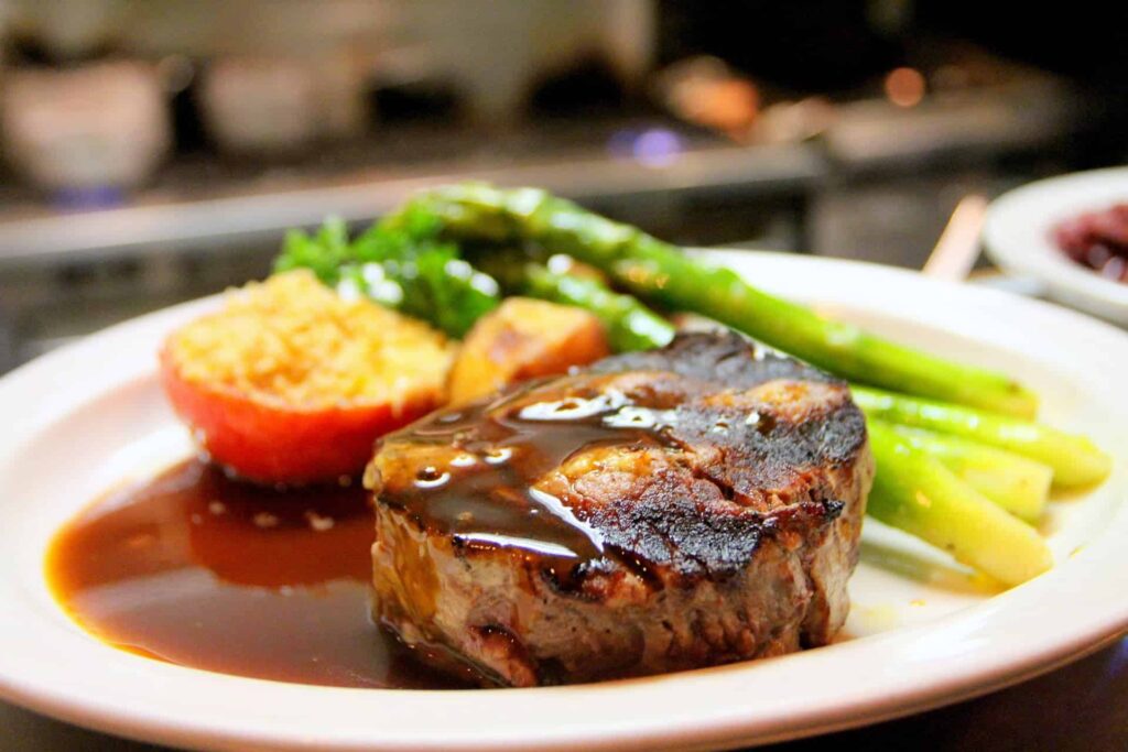 Plate with a medium-cooked steak topped with glaze, served with asparagus, roasted potato, and a brown sauce in a bright dining setting