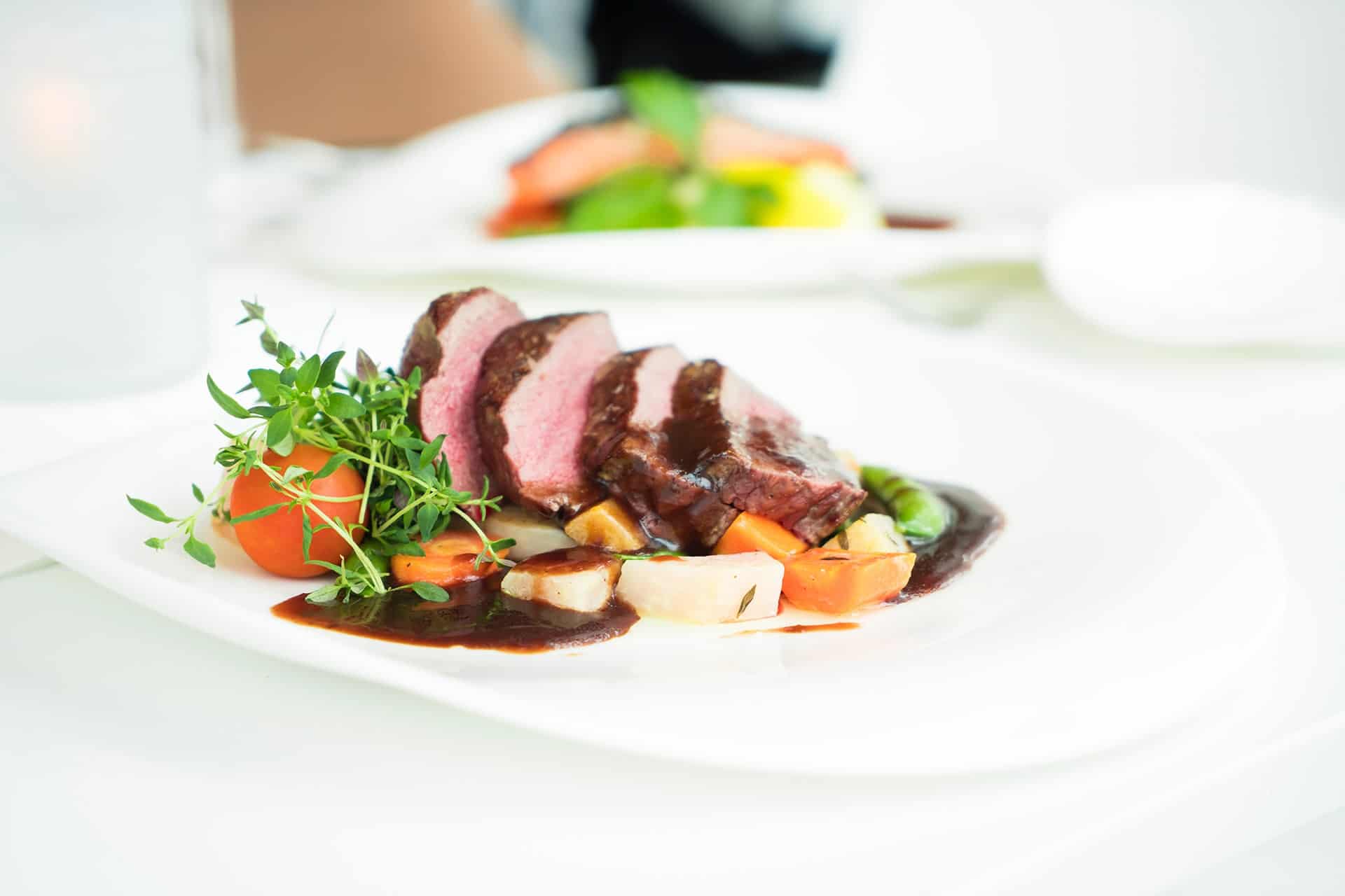 pexels-terje-sollie-299347 - RIM Digital Sliced medium-rare beef with dark glaze on a bed of diced carrots and potatoes, garnished with fresh greens on a white plate.