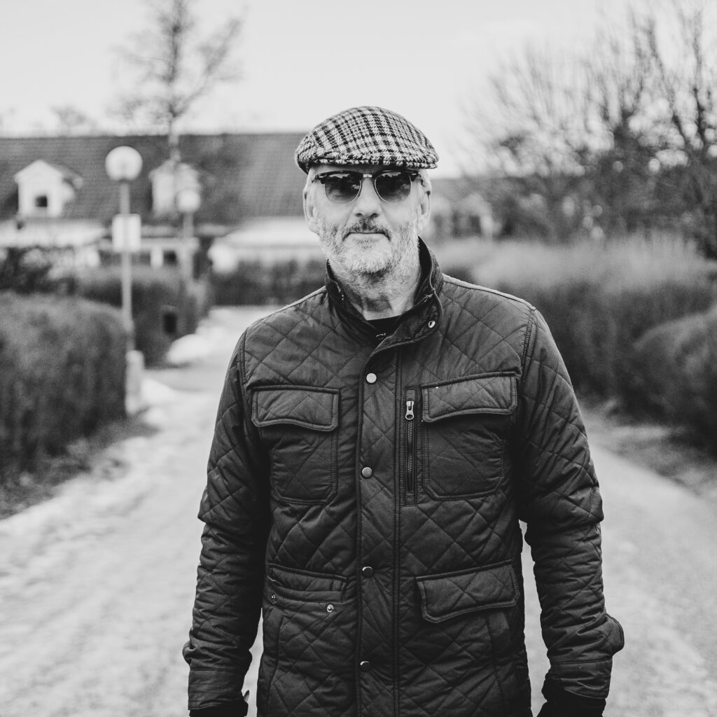 Jens Andersson 2024-02-04 - RIM Digital Jens Andersson i Enköping som står på en väg med bostadshus och träd i bakgrunden. Jens har grått skägg och bär solglasögon, en brun och svart rutig keps, och en olivgrön quiltad jacka med ståkrage och flera fickor. Bakom honom ser det ut att vara sent på hösten eller tidigt på våren eftersom träden är kala och det finns lite snö kvar på marken. Mannen verkar vara avslappnad och är fotograferad i dagsljus.