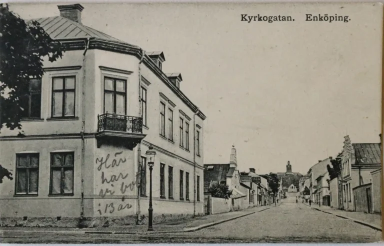 Kyrkogatan i Enköping omkring 1900–1910, visande kullerstensgata kantad av låg bebyggelse, några gående personer och enklare trädplantering.